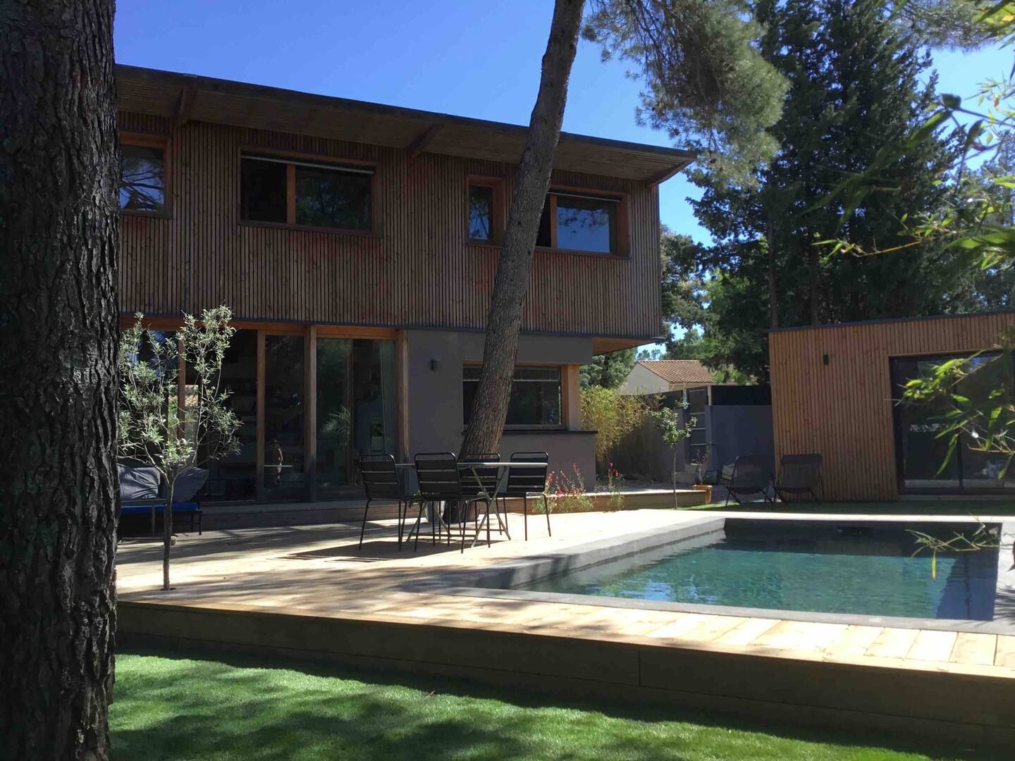 La maison en bois, sa terrasse et la piscine sous les pins